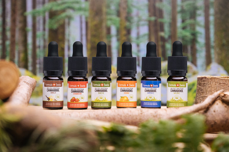 Seis óleos de CBD de espetro completo da Formula Swiss com diferentes óleos de base e sabores, em frente a uma floresta