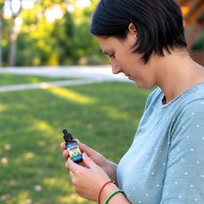 Mulher num parque a ler o rótulo de um frasco de óleo de CBD, com t-shirt azul às bolinhas e pulseiras coloridas